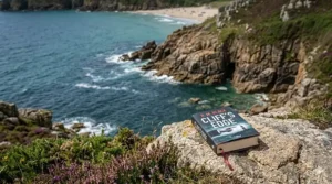 A detailed close-up of a crime thriller paperback resting on a granite cliff ledge overlooking a dramatic Cornish cove under natural daylight.
