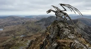 A massive skeletal dragon perched on a rocky mountain peak overlooking a vast, rugged valley with a small stone ruin in the distance under a grey sky.